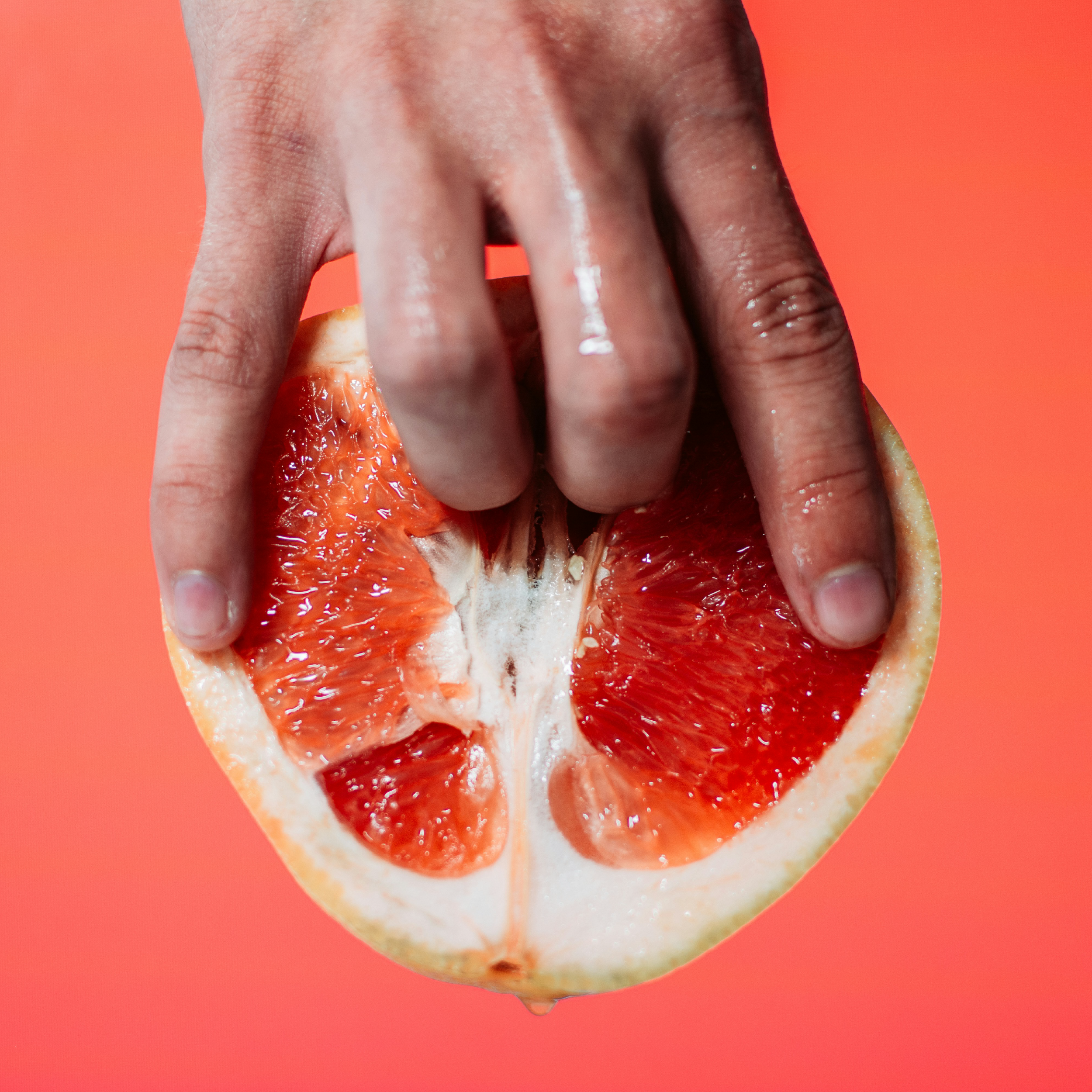 Close-up of a hand gently pressing into a halved grapefruit, artistically symbolizing female sensuality and pleasure, with a bright coral background.