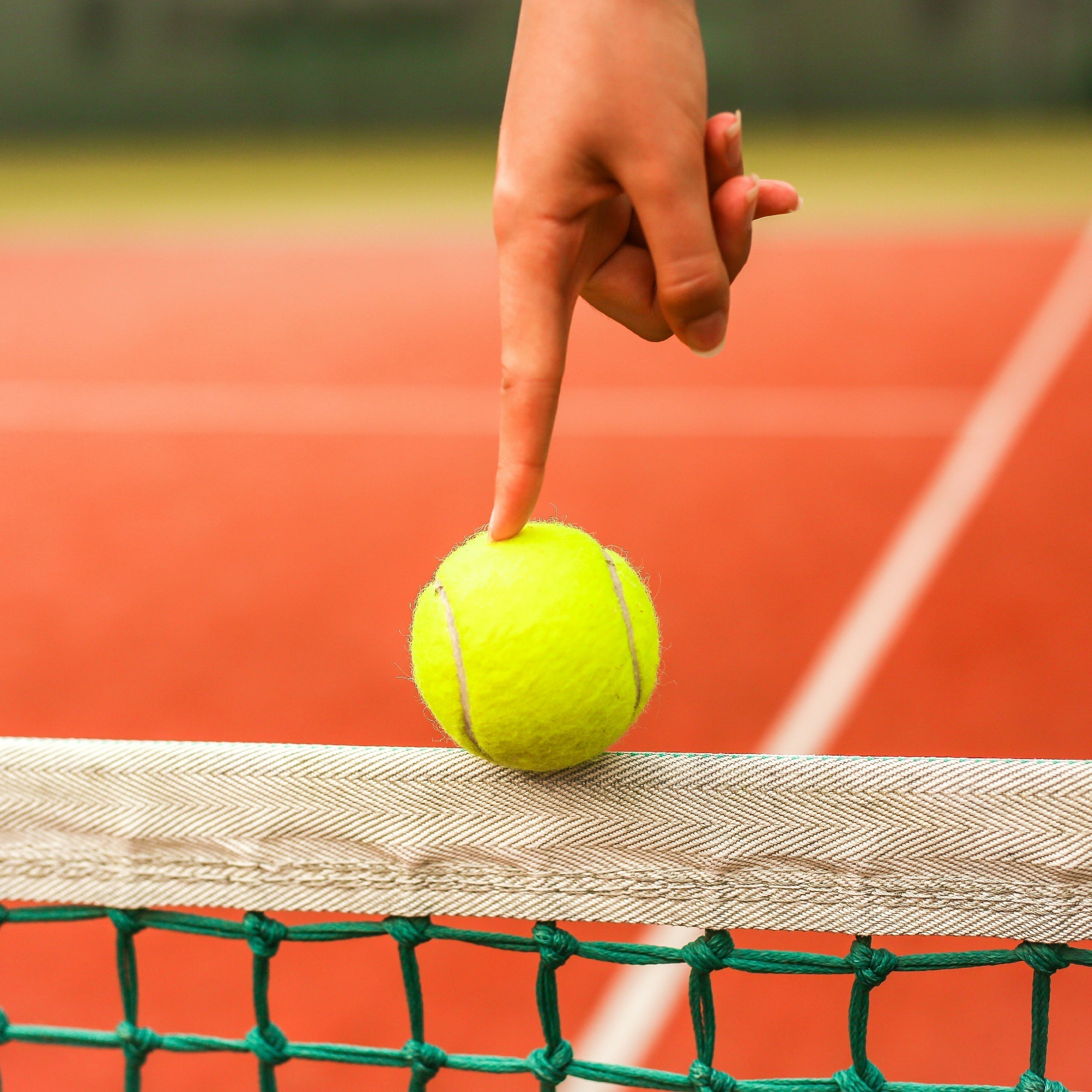 Close-up of a hand gently pressing a tennis ball balanced on a net, with a clay court in the background.