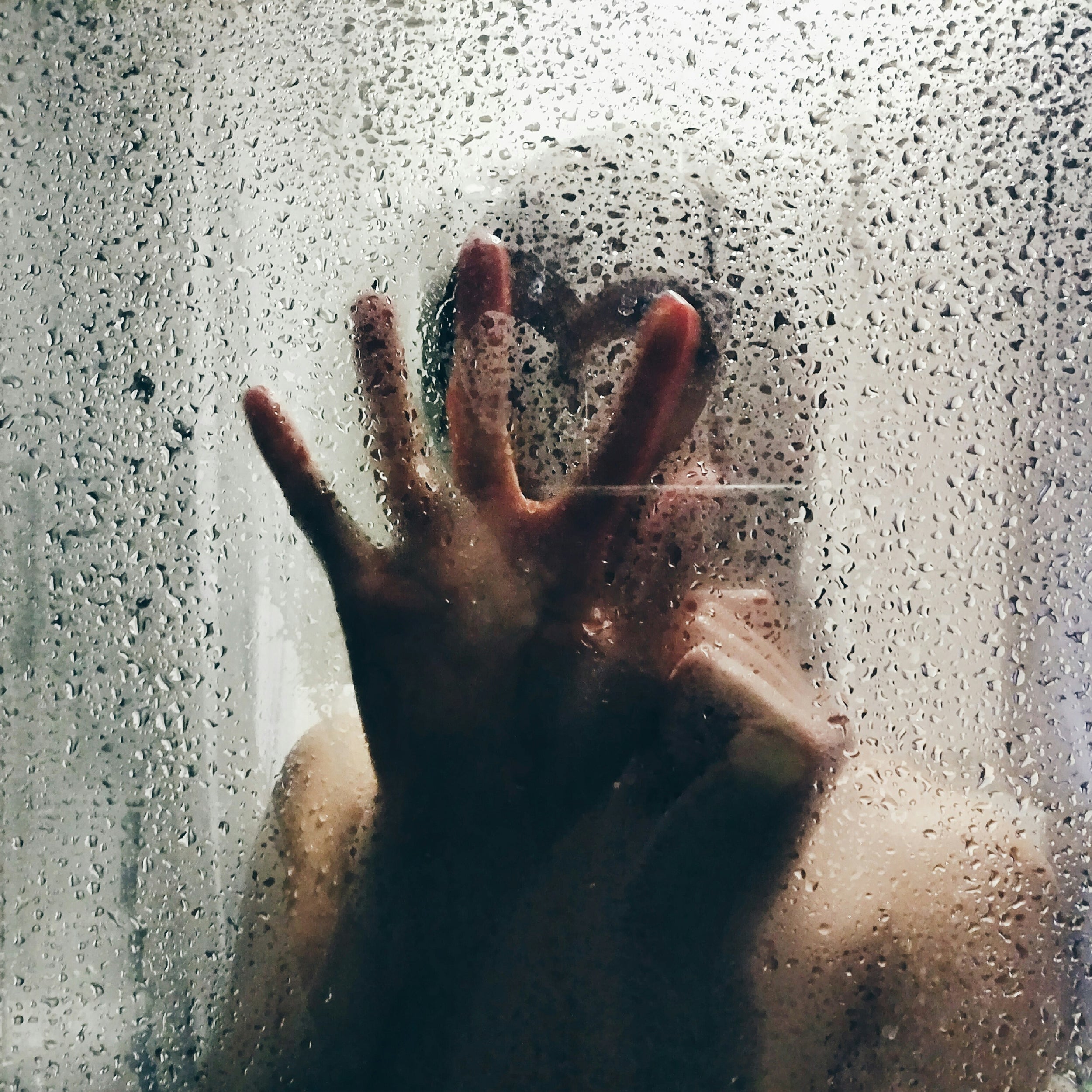 A blurred silhouette of a person behind a fogged glass shower door with their hand pressed against it, evoking a sense of emotion or vulnerability.