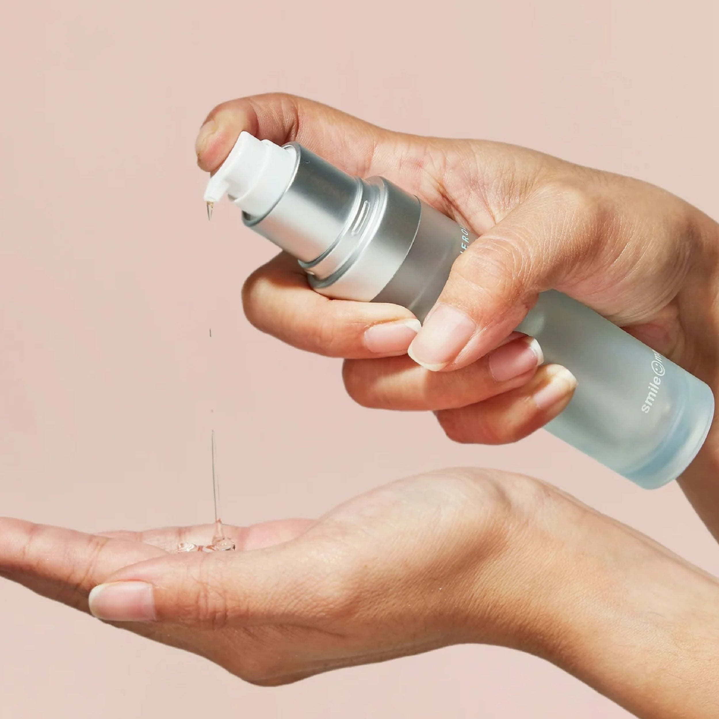 Hands applying clear lubricant from a silver pump bottle against a soft pink background.