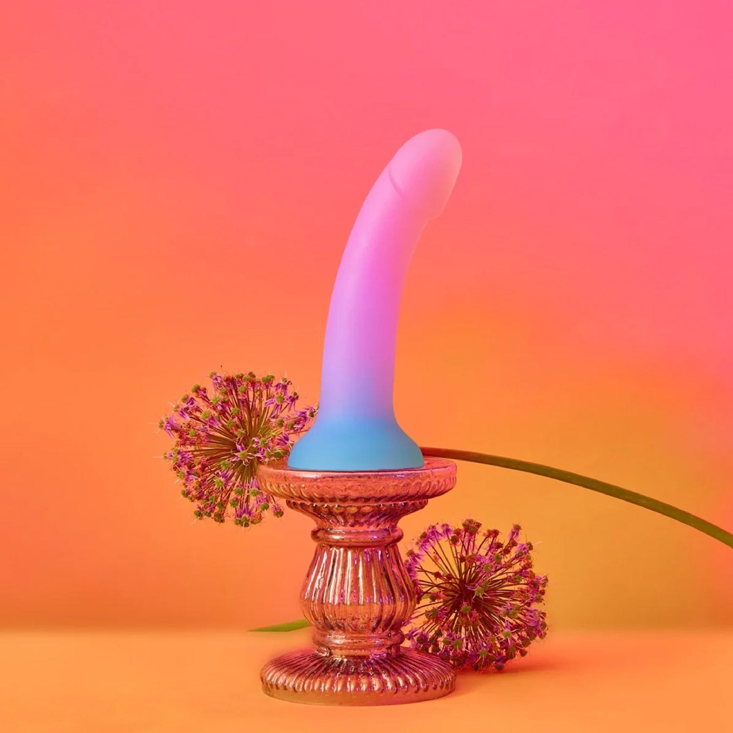 Curved silicone dildo in a gradient of pink to blue, displayed on an ornate stand with floral accents against a vibrant orange-pink background.