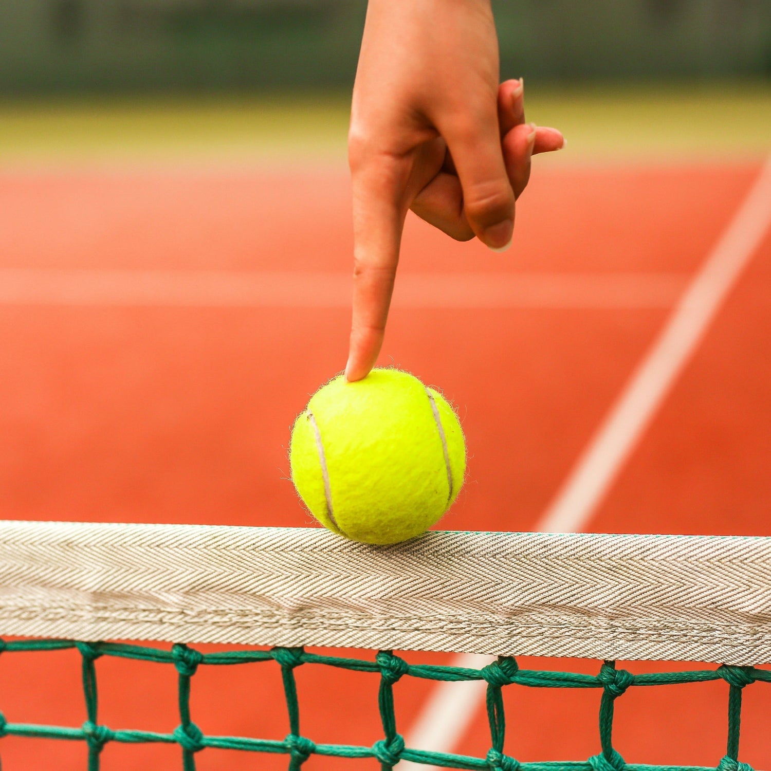 Close-up of a hand gently pressing a tennis ball balanced on a net, with a clay court in the background.